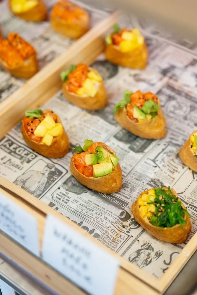 Inari rellenos de salmón con aguacate y mango, y uno de maíz con wakame, sobre papel de periódico en su bandeja de madera